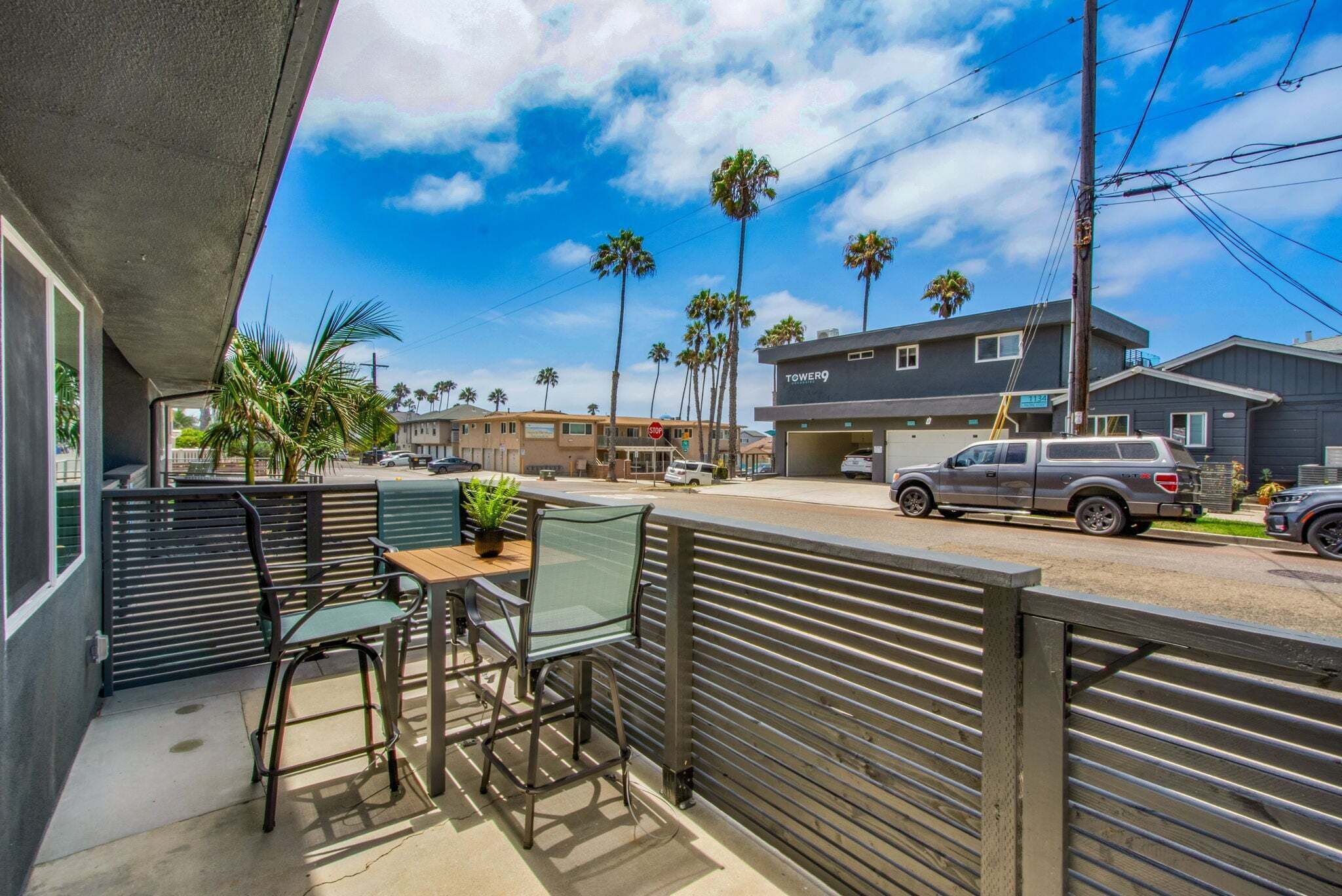 A balcony with a table and chairs, overlooking a street with palm trees and a building labeled 'TOWER 9'.