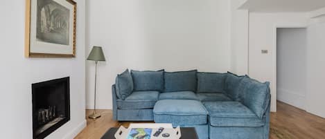 A living room with a blue sectional sofa, a fireplace, a floor lamp, and a coffee table with a magazine and remote controls.