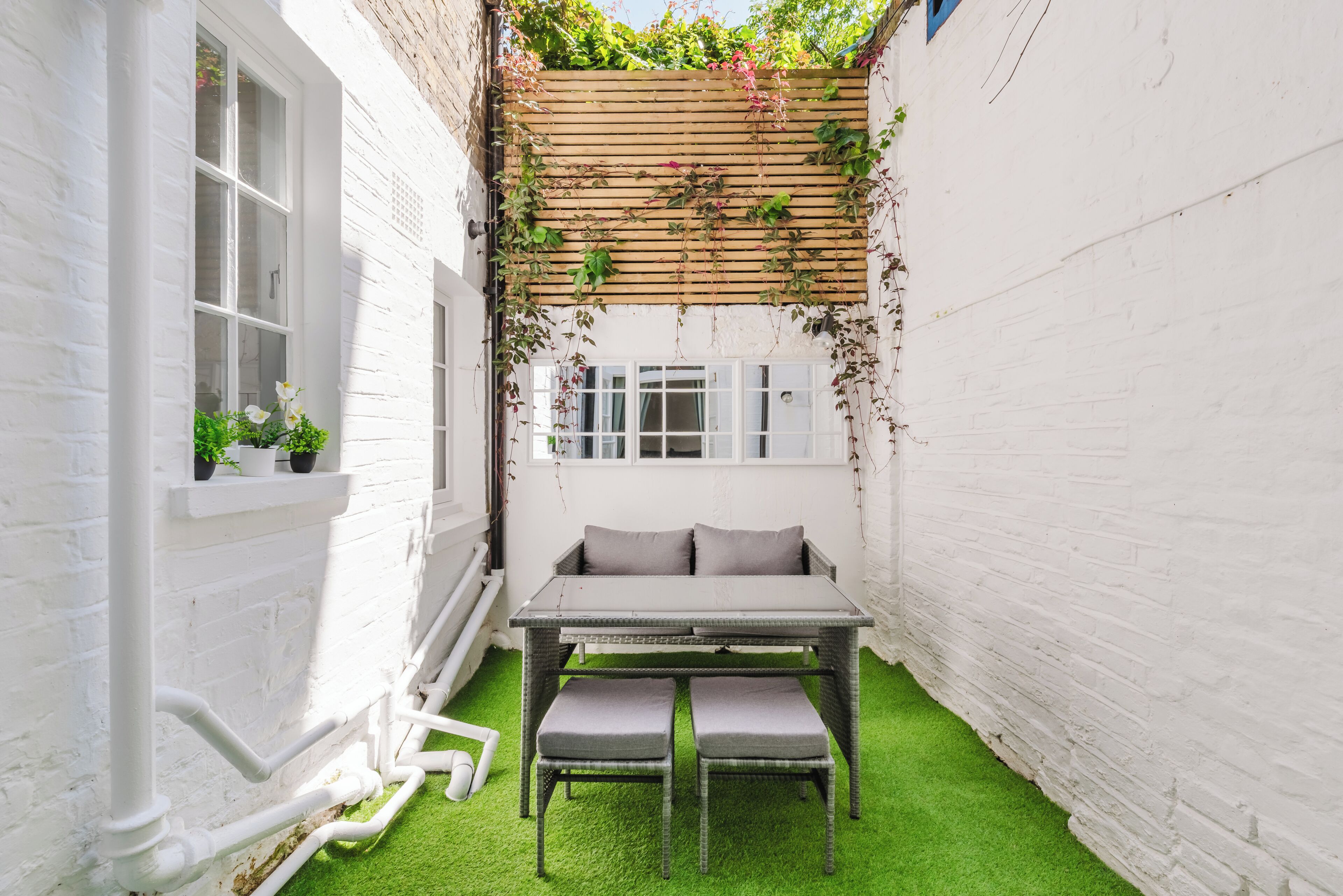 A small outdoor seating area with a table and benches, surrounded by white brick walls and a wooden trellis with climbing plants.