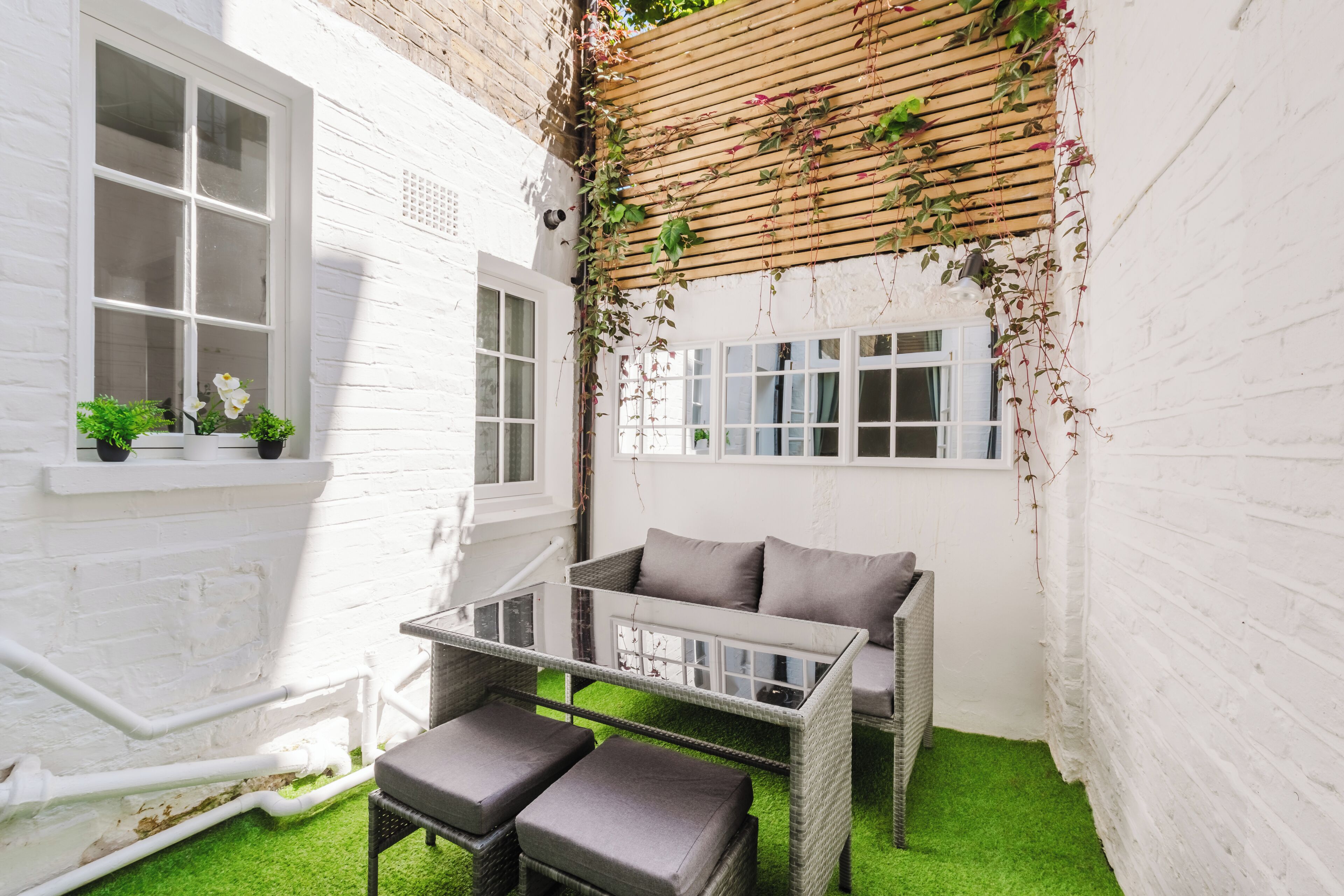 A white brick room with a glass table and two grey cushioned chairs, a wooden slatted ceiling, and a window with white shutters.