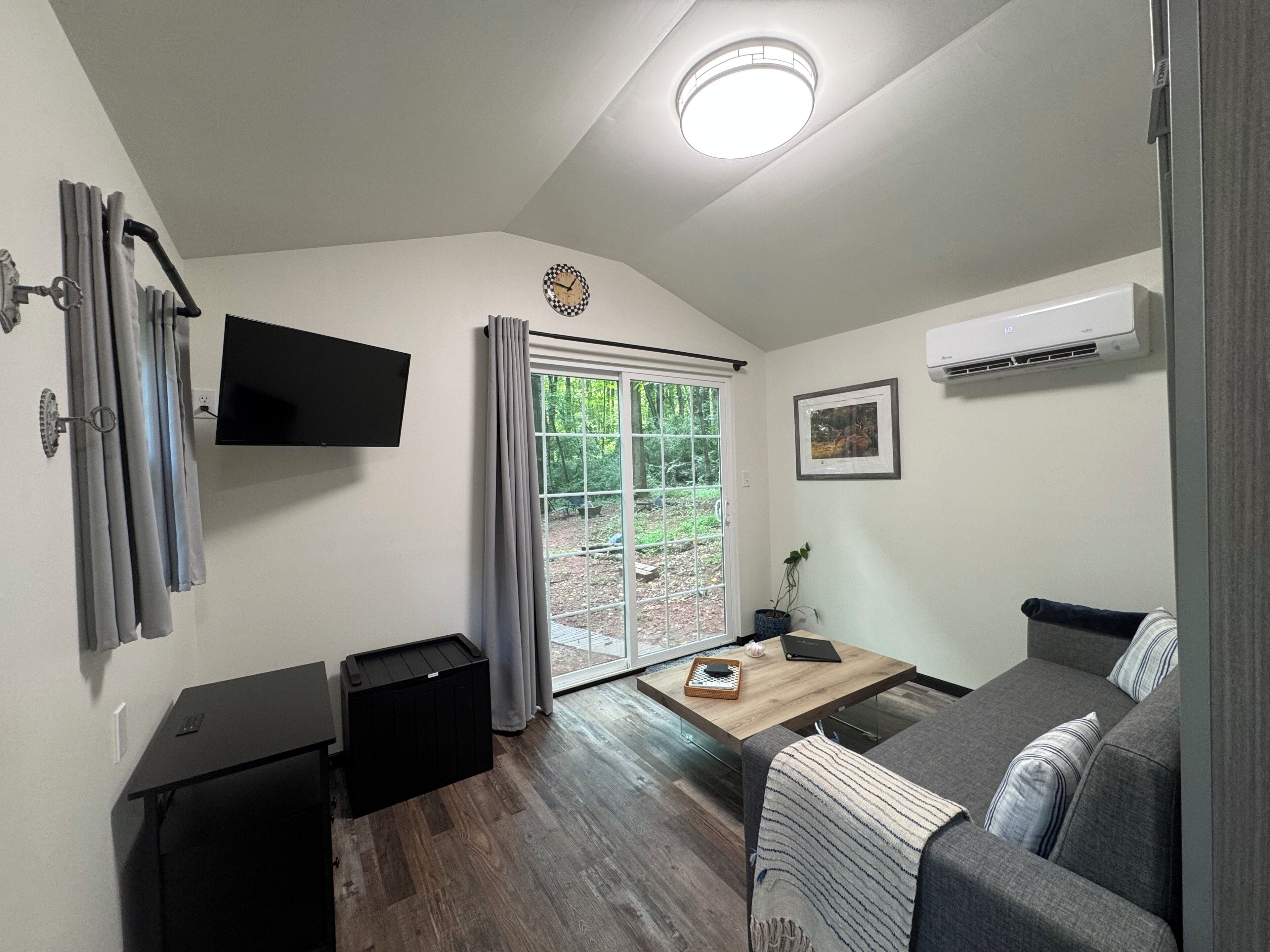 A compact living space with a sofa, a small table, a TV mounted on the wall, and a window with curtains.