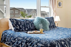 A bedroom with a bed, a fluffy pillow, a lamp, and a window with a view of houses. - Waterfront - Sauna -steps To Beach! (Saco)