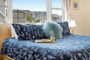 Un dormitorio con cama, almohada mullida, lámpara y ventana con vista a casas.