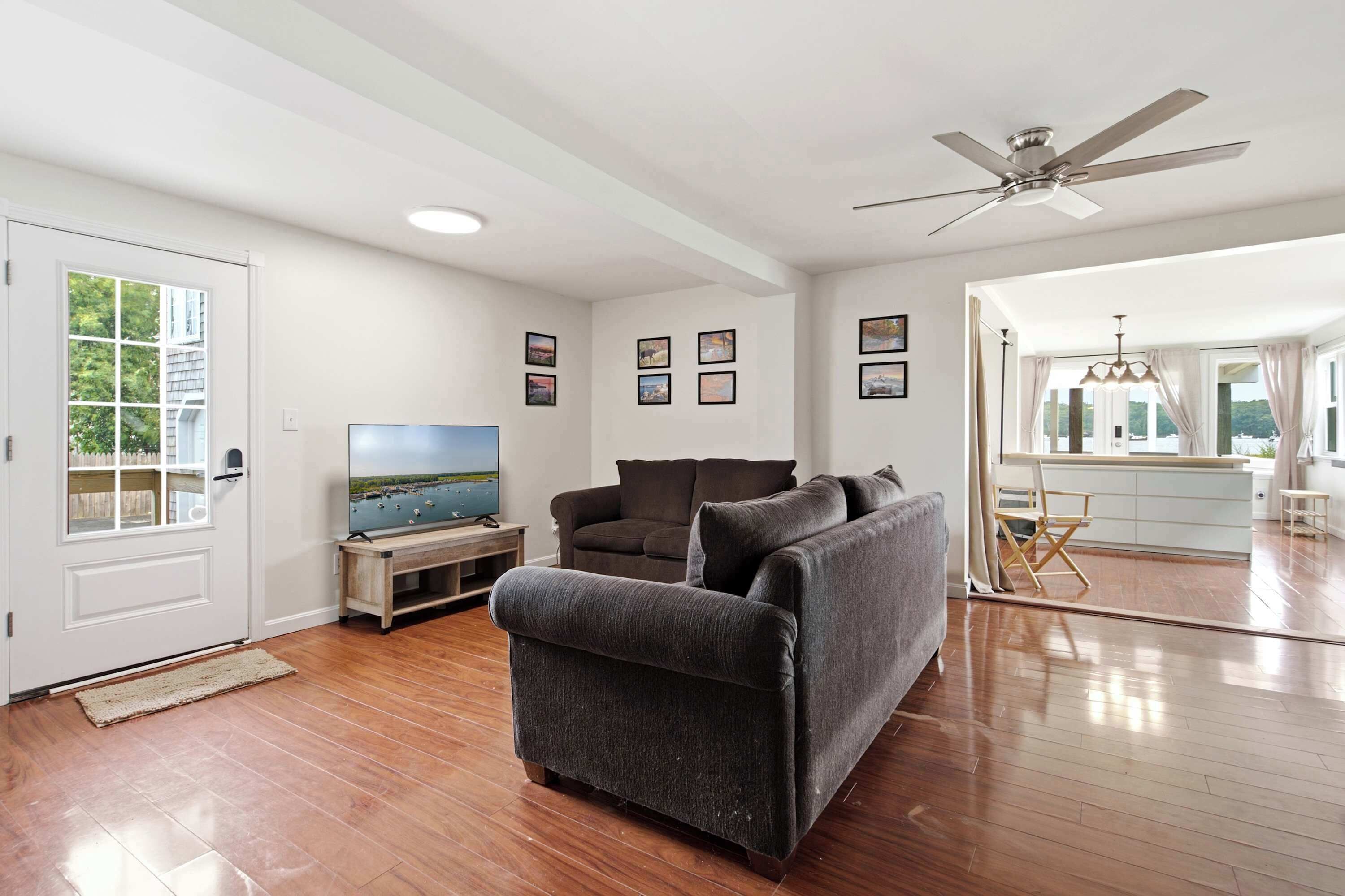 A spacious living room with a flat-screen TV, a grey sofa, wooden flooring, and a ceiling fan.