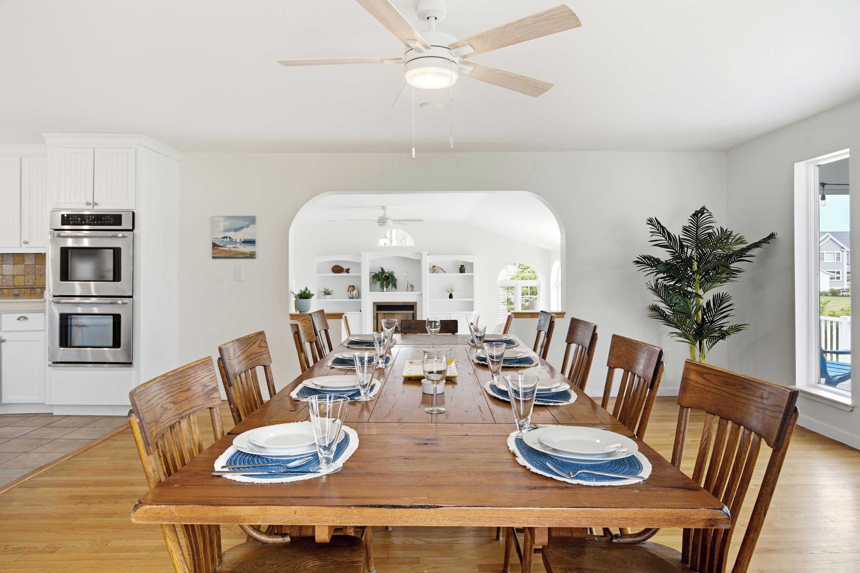 A dining area with a wooden table set for six, surrounded by wooden chairs, in a modern kitchen with white cabinets and a ceiling fan.