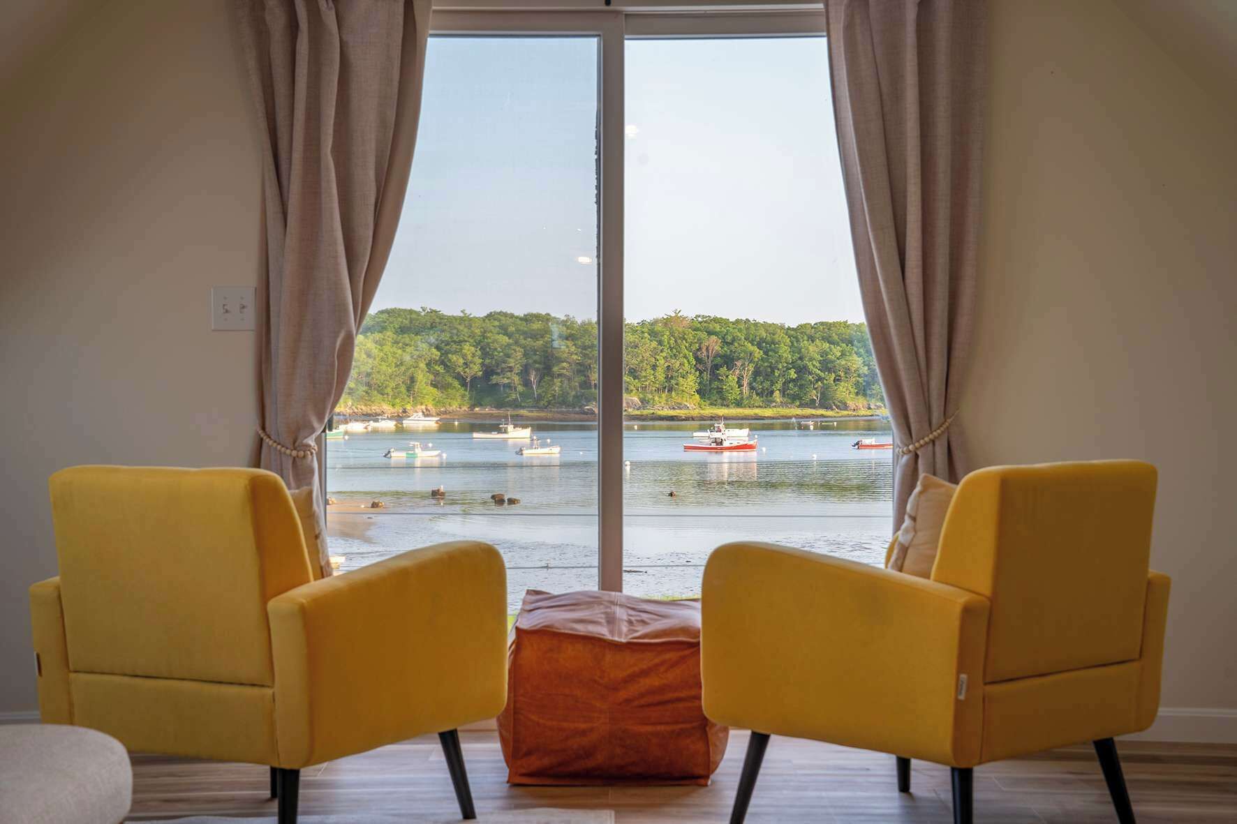 A room with a large window, two yellow chairs, and a small orange ottoman.