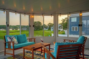 Un solarium con vistas a un paisaje verde y una casa azul. Hay una mesa de madera con una copa de vino tinto.