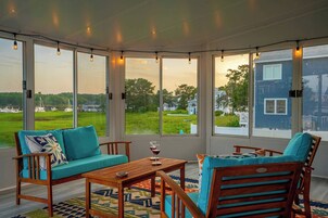 Un solarium con vistas a un paisaje verde y una casa azul. Hay una mesa de madera con una copa de vino tinto.