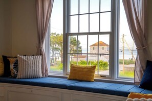 A room with a large window, a blue cushioned bench, and a view of a house and trees. - Waterfront - Sauna -steps To Beach! (Saco)