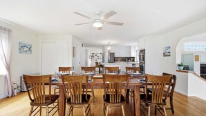 A dining area with a wooden table and chairs, a ceiling fan, and a kitchen in the background. - Waterfront - Sauna -steps To Beach! (Saco)