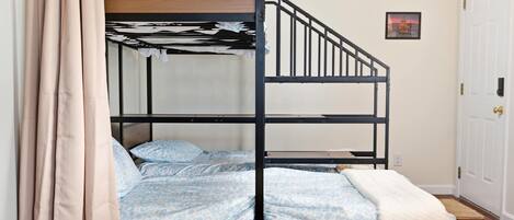 Une chambre avec un lit superposé doté d’une tête de lit en bois, un lit simple avec une literie à motifs et une porte blanche ornée d’un petit tableau au mur.