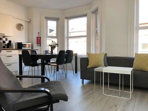 A modern living room with a sofa, a dining table with chairs, a small white table, and a kitchen area with a microwave. - Spacious 3-Bed Apartment in Queen's Quarter (Belfast)