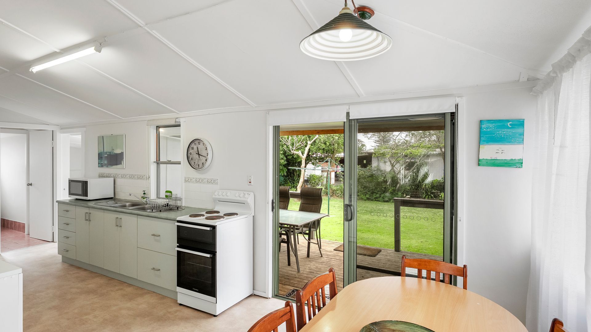 A modern kitchen with a dining area, a clock on the wall, and a view of the garden through the glass door.