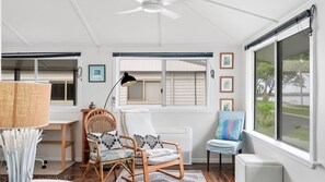 A room with a ceiling fan, a window with blinds, a wicker chair, a desk with a lamp, and framed pictures on the wall.