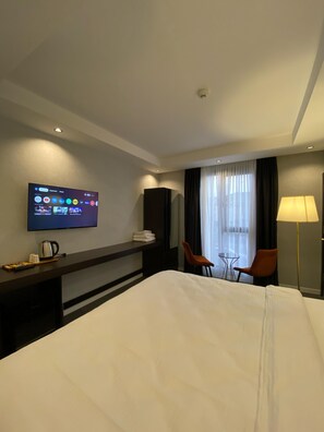 Een hotelkamer met een bed, een aan de muur bevestigde televisie, een bureau met een waterkoker, twee stoelen, een lamp en een raam met gordijnen.