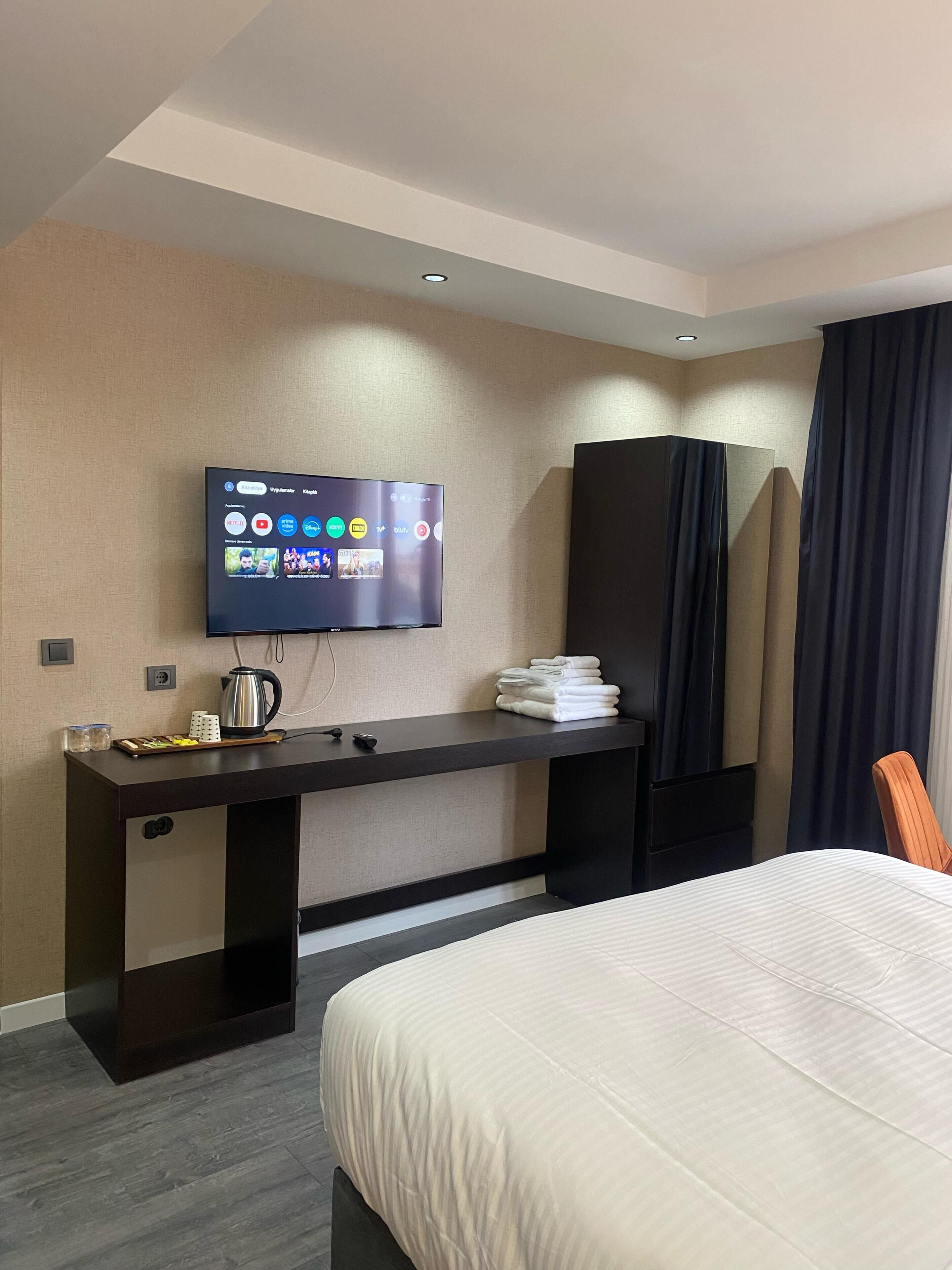 Hotelkamer met een bed, een bureau met een flatscreen-tv waarop streamingdiensten worden afgespeeld, een waterkoker en een stapel handdoeken.