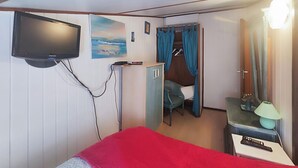 Una habitación con cama, televisión montada en la pared, un mueble pequeño, escritorio con lámpara y puerta que da a un baño.