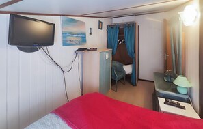 Ein Zimmer mit einem Bett, einem an der Wand befestigten Fernseher, einer kleinen Kommode, einem Schreibtisch mit Lampe und einer Tür zu einem Badezimmer.