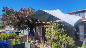 Un espacio al aire libre techado con una mesa y sillas, rodeado de plantas en macetas y un árbol con flores moradas.