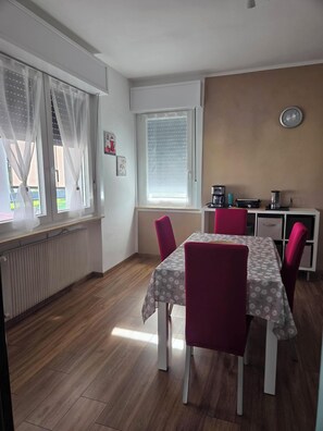 Un comedor con una mesa lista para cuatro personas, una zona de cocina con electrodomésticos y un ventanal con cortinas.