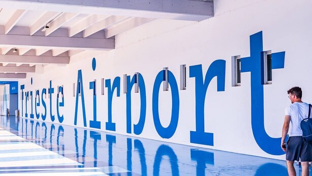 A modern airport interior with large signage reading 'Trieste Airport' and a person standing near the entrance.