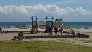Una playa con un parque infantil, que incluye columpios y un tobogán, y una zona arenosa con gente.