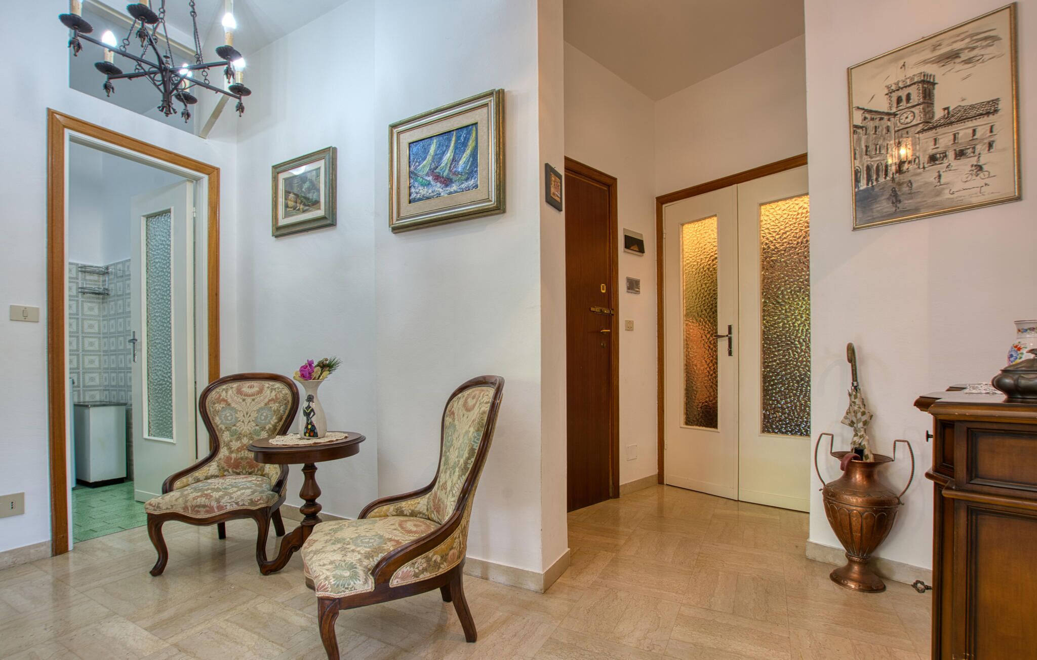 Un couloir avec deux fauteuils rembourrés, une petite table, un vase de fleurs et une porte dont les panneaux sont en verre dépoli.