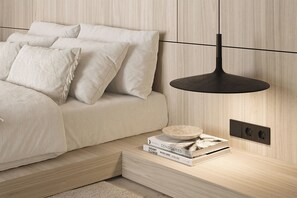 Ein modernes Schlafzimmer mit einem Bett, Kissen, einem Nachttisch mit Büchern, einer Wandleuchte und Steckdosen.