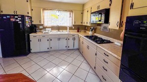 Une cuisine avec des meubles blancs, un réfrigérateur noir, une cuisinière et une fenêtre avec des stores.