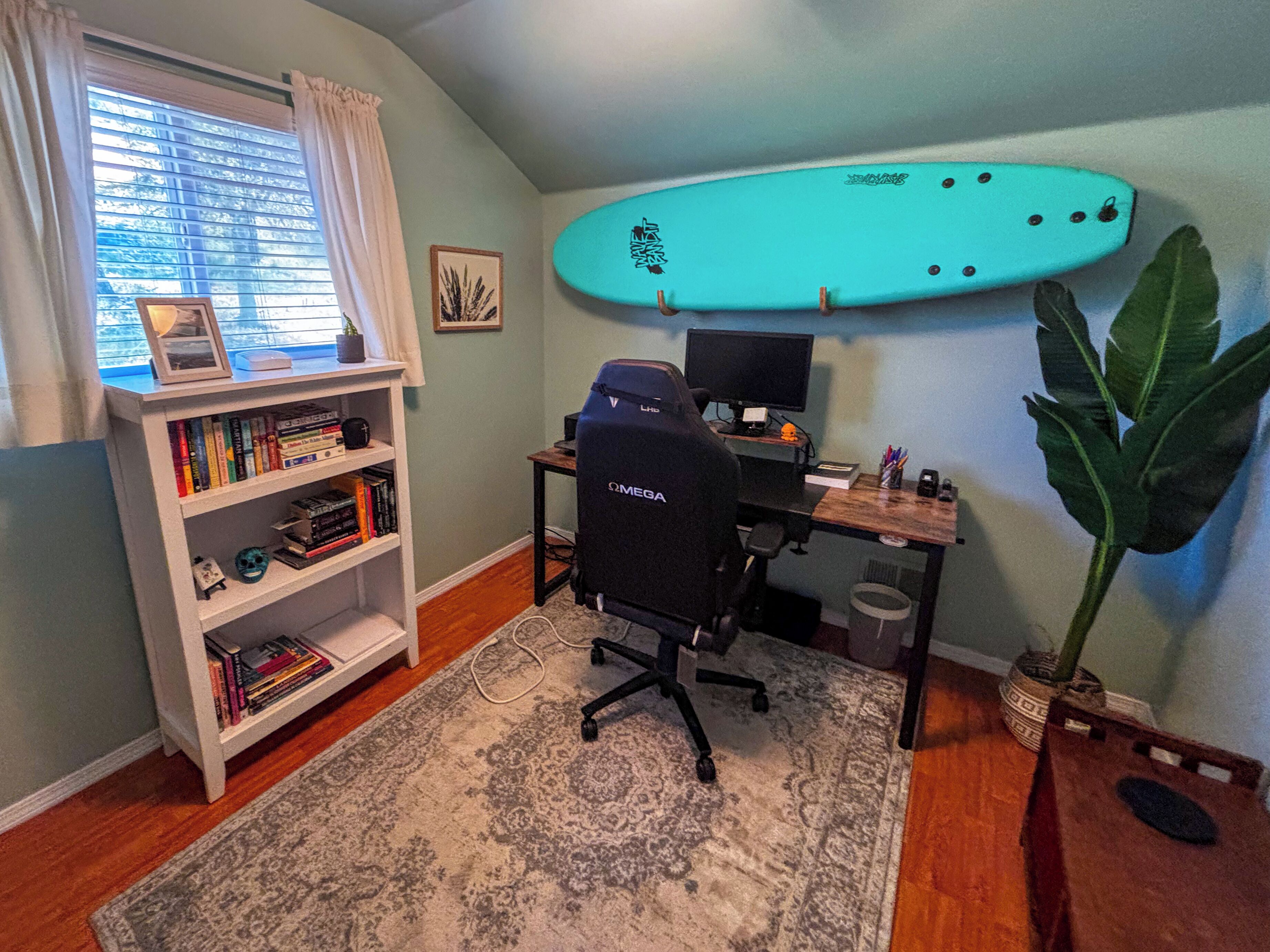 Ein Zimmer mit Schreibtisch, Computer, Bücherregal, Surfbrett und einer großen Pflanze.