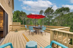 Eine Holzterrasse mit Tisch und Stühlen, einem roten Sonnenschirm und Blick auf Bäume.