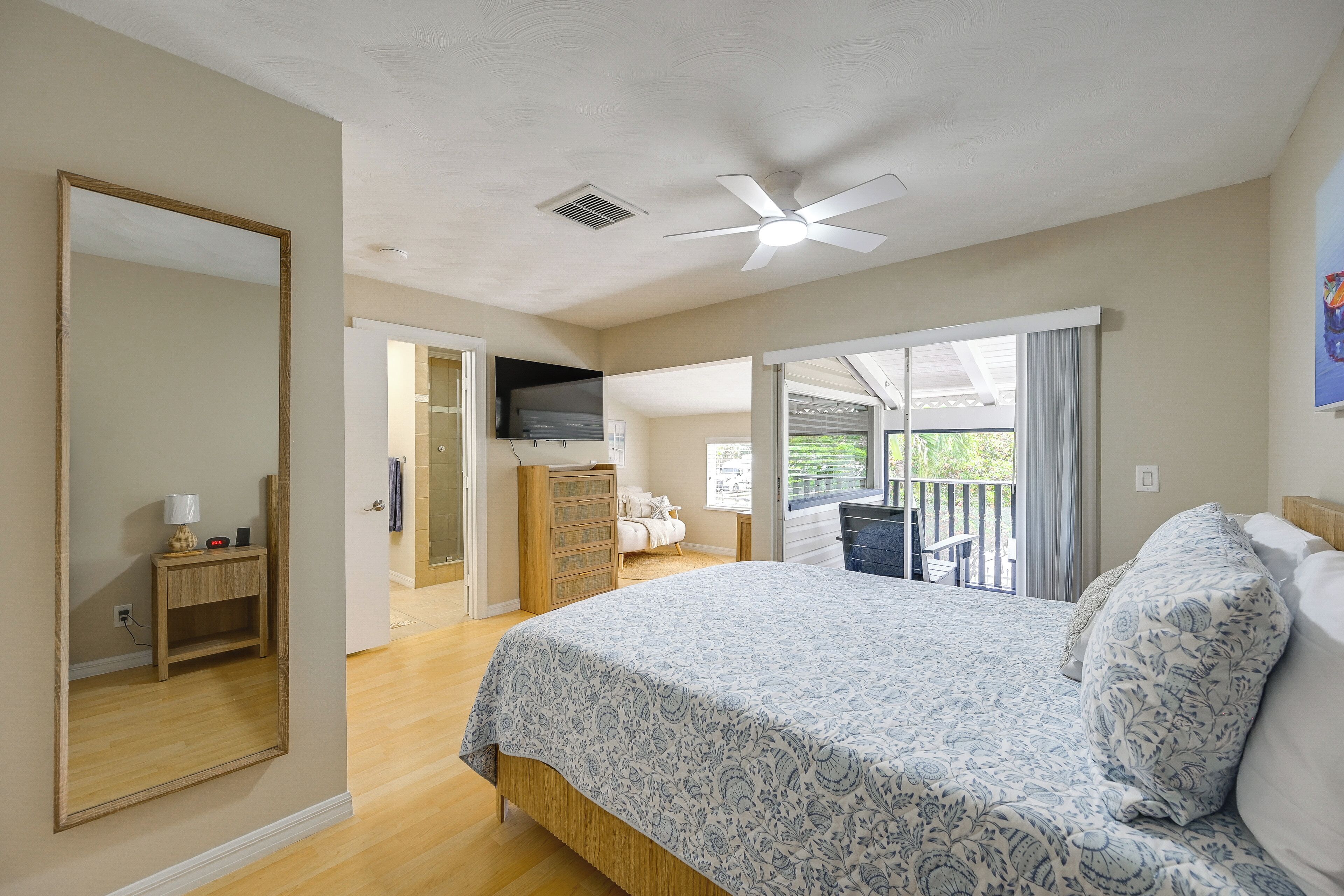 A bedroom with a large bed, a ceiling fan, a TV, and a balcony with a view.
