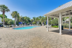 Ein Pool mit sechseckigen Fliesen, umgeben von Liegestühlen und Sonnenschirmen, mit Palmen und einer überdachten Sitzgelegenheit im Hintergrund.