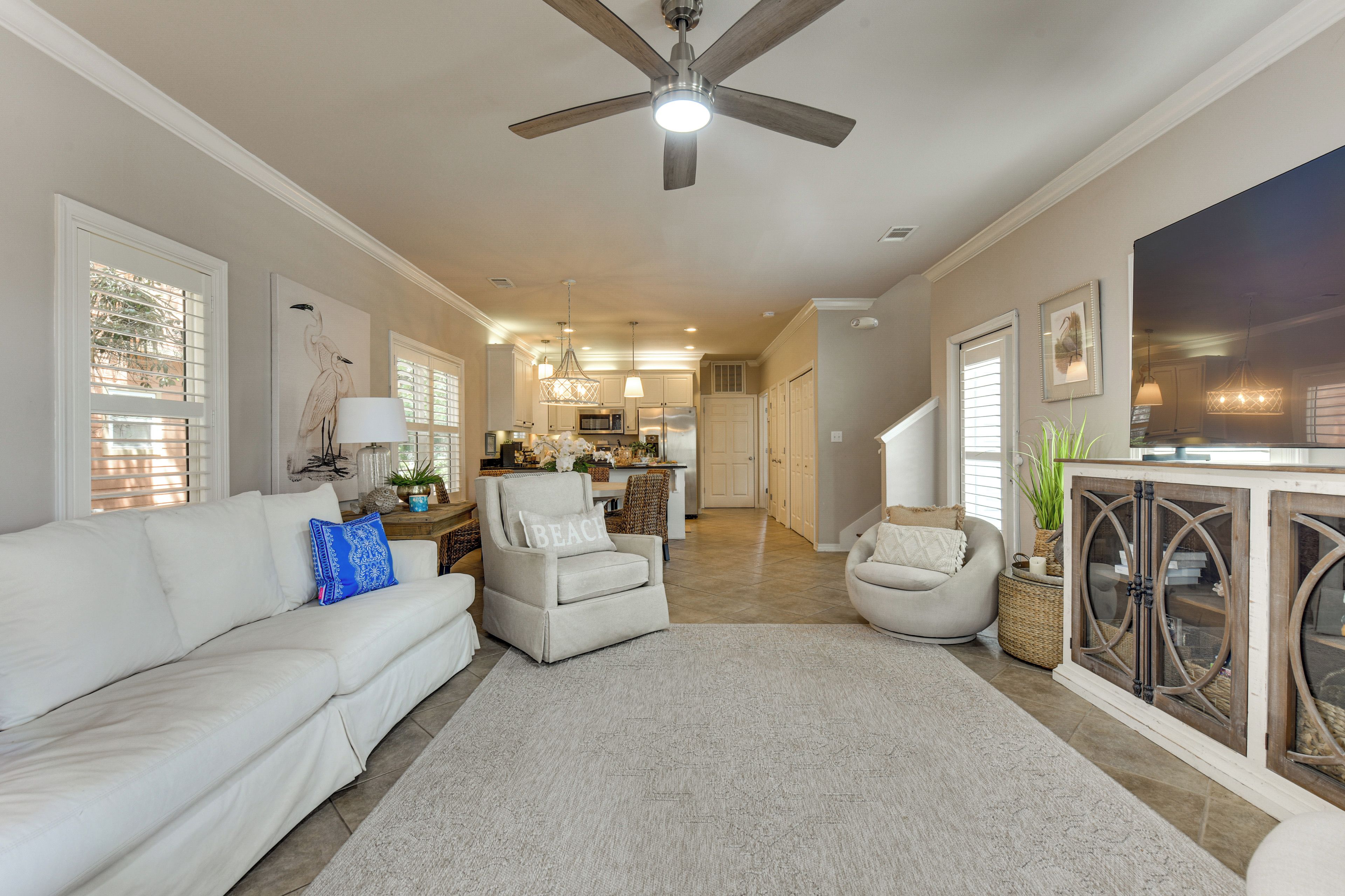 A spacious living room with a white sofa, two armchairs, a ceiling fan, and a view into the kitchen.