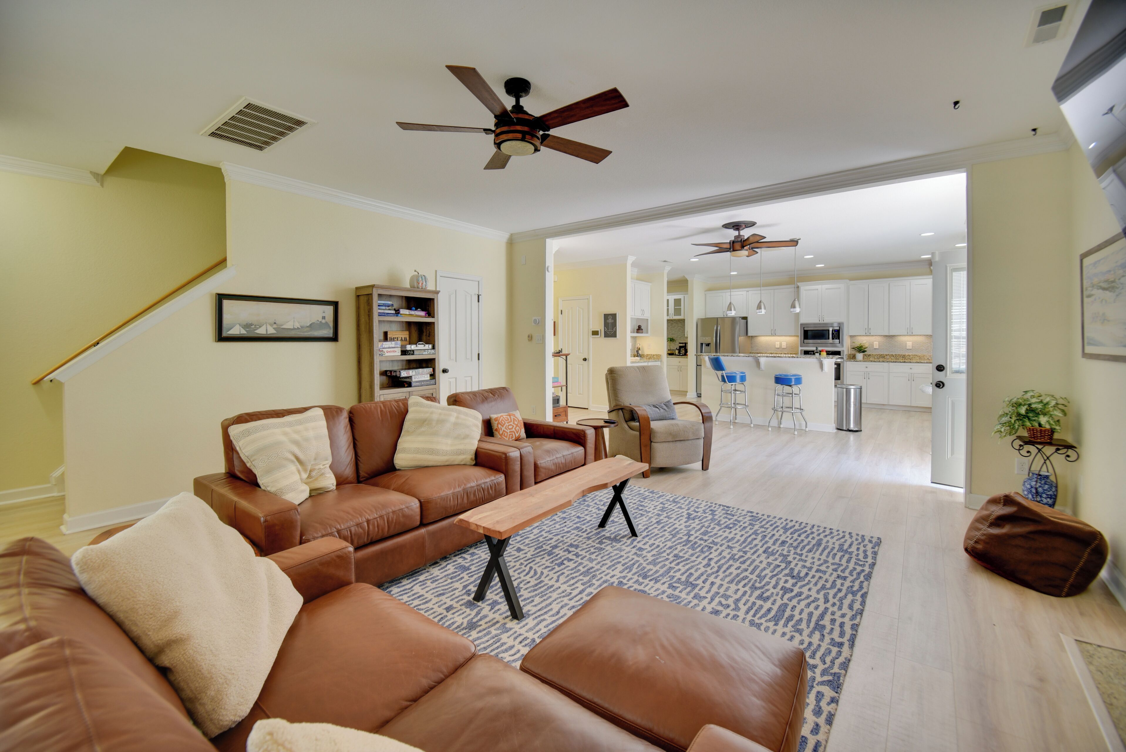 A spacious living room with a brown leather sofa, a wooden coffee table, and a patterned rug. There is a ceiling fan, a staircase, and a view into the kitchen area.