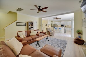 Ein geräumiges Wohnzimmer mit einem braunen Ledersofa, einem Holztisch und einem gemusterten Teppich. Es gibt einen Deckenventilator, eine Treppe und einen Blick in den Küchenbereich.