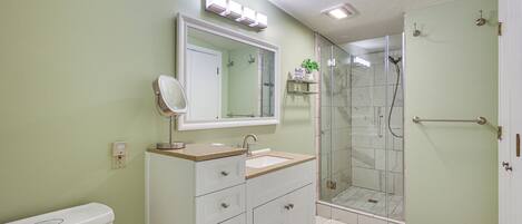 Un baño con inodoro, lavamanos con espejo, ducha con puerta de vidrio y estante con un letrero.