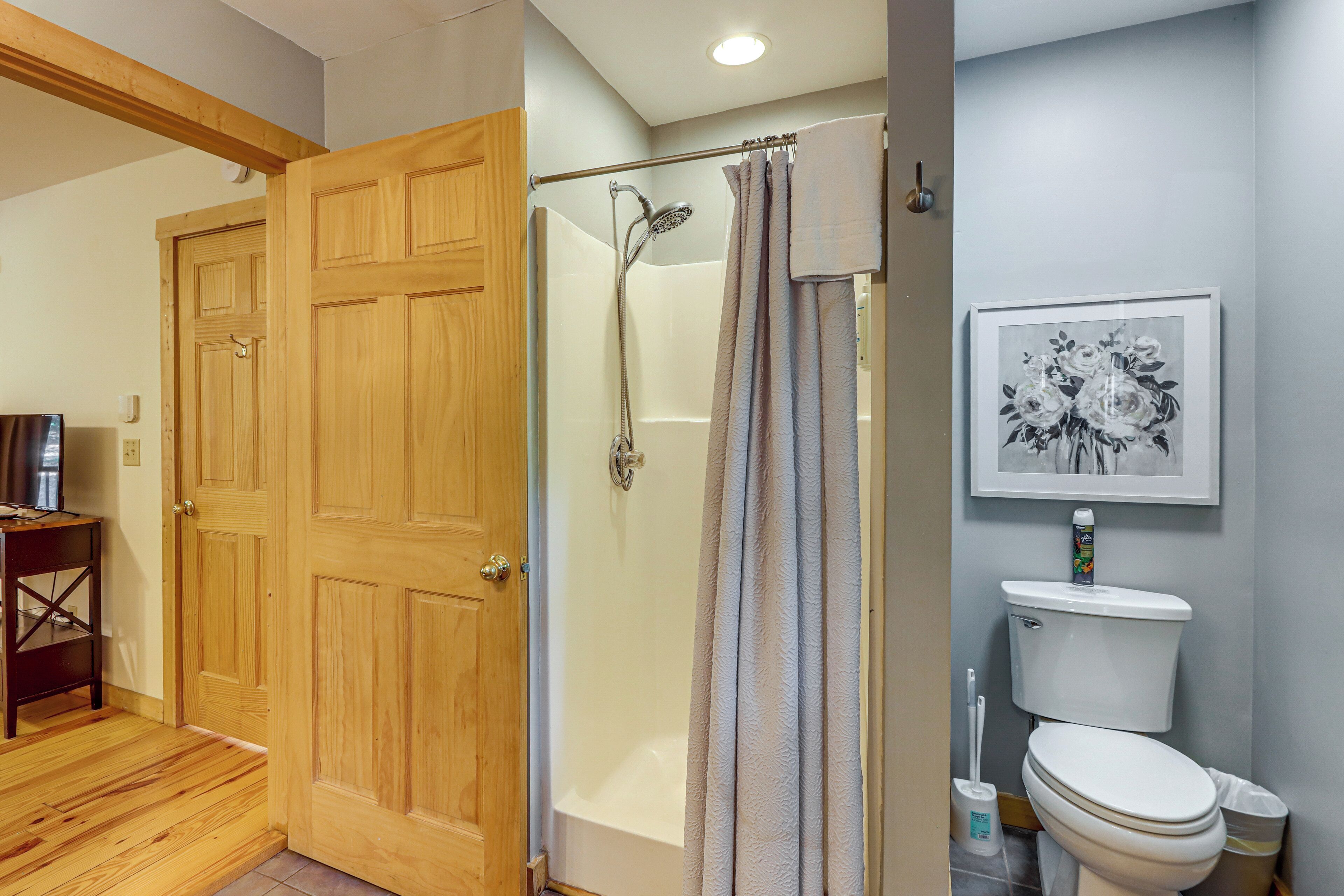 A wooden door, a shower area with a glass door, and a toilet with a framed floral print on the wall.