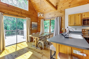 Interno di una baita in legno con zona pranzo, cucina e ampie finestre che si affacciano su una foresta.