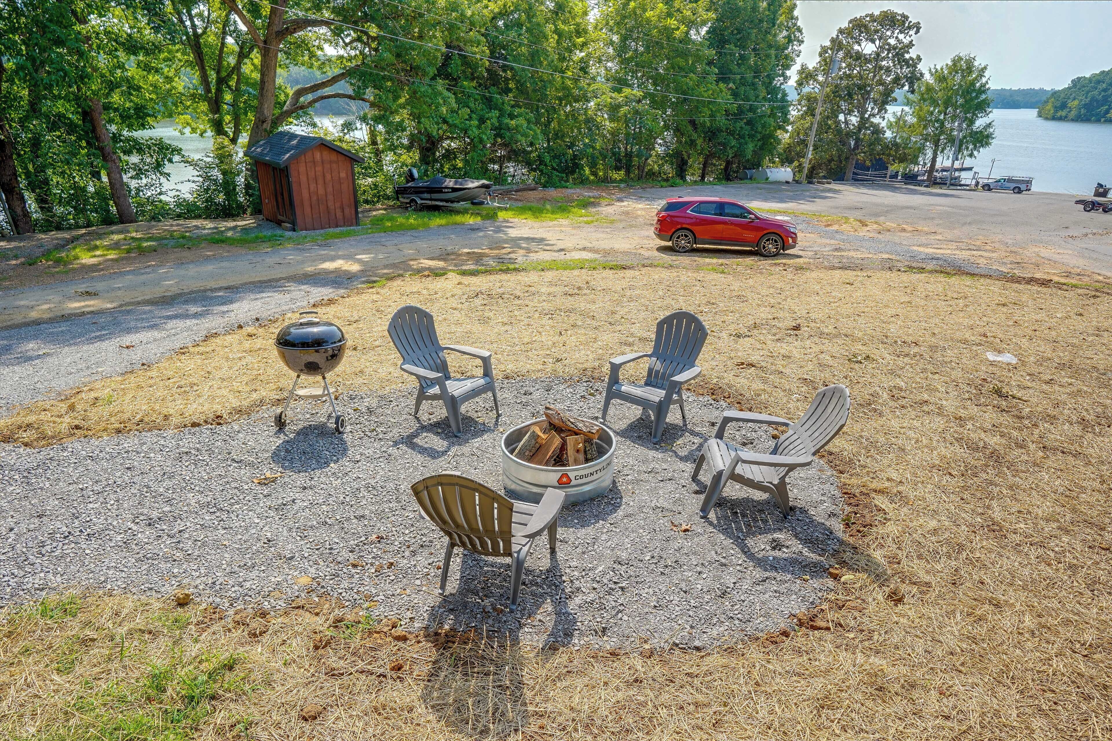 Een grindgebied met terras, een vuurkorf en uitzicht op een meer met boten en een rode auto die in de buurt geparkeerd staat.