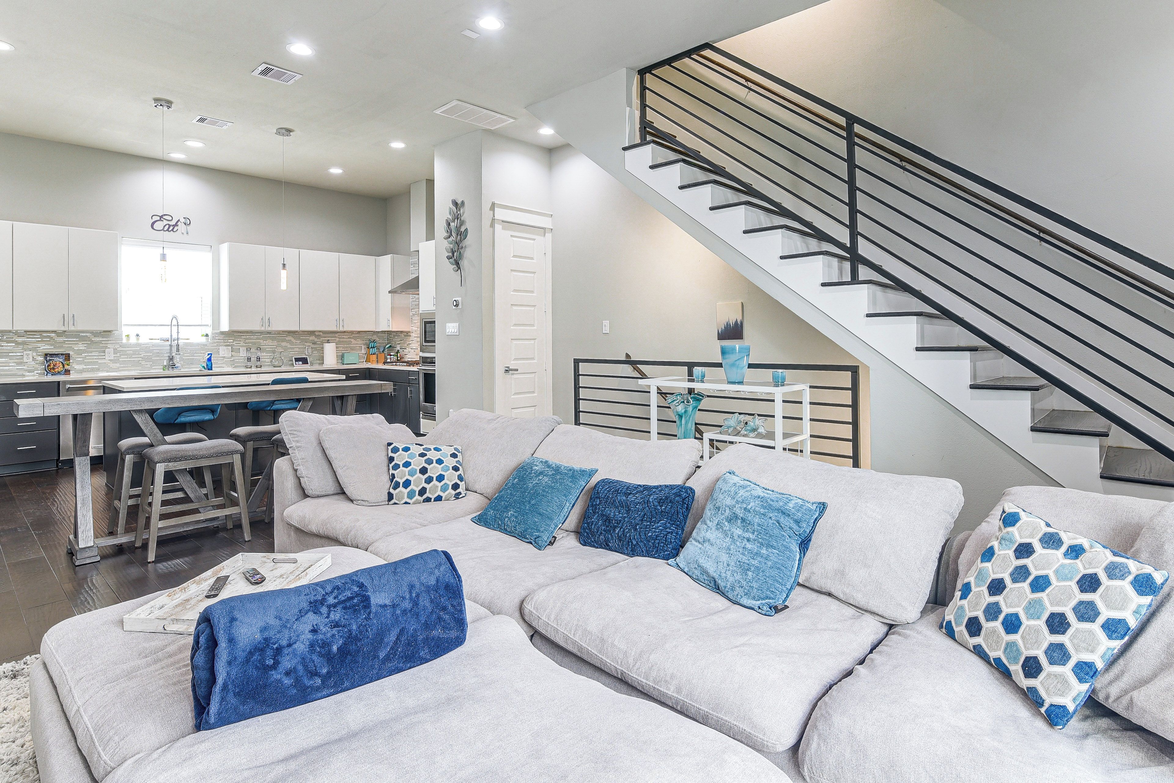 A modern living room with a grey sofa, a kitchen in the background, and a staircase.