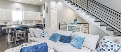 A modern living room with a grey sofa, a kitchen in the background, and a staircase.