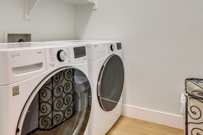 A white LG ThinQ front-loading washing machine with a control panel, a transparent door with a black swirl pattern, and a white wall with a wooden floor.
