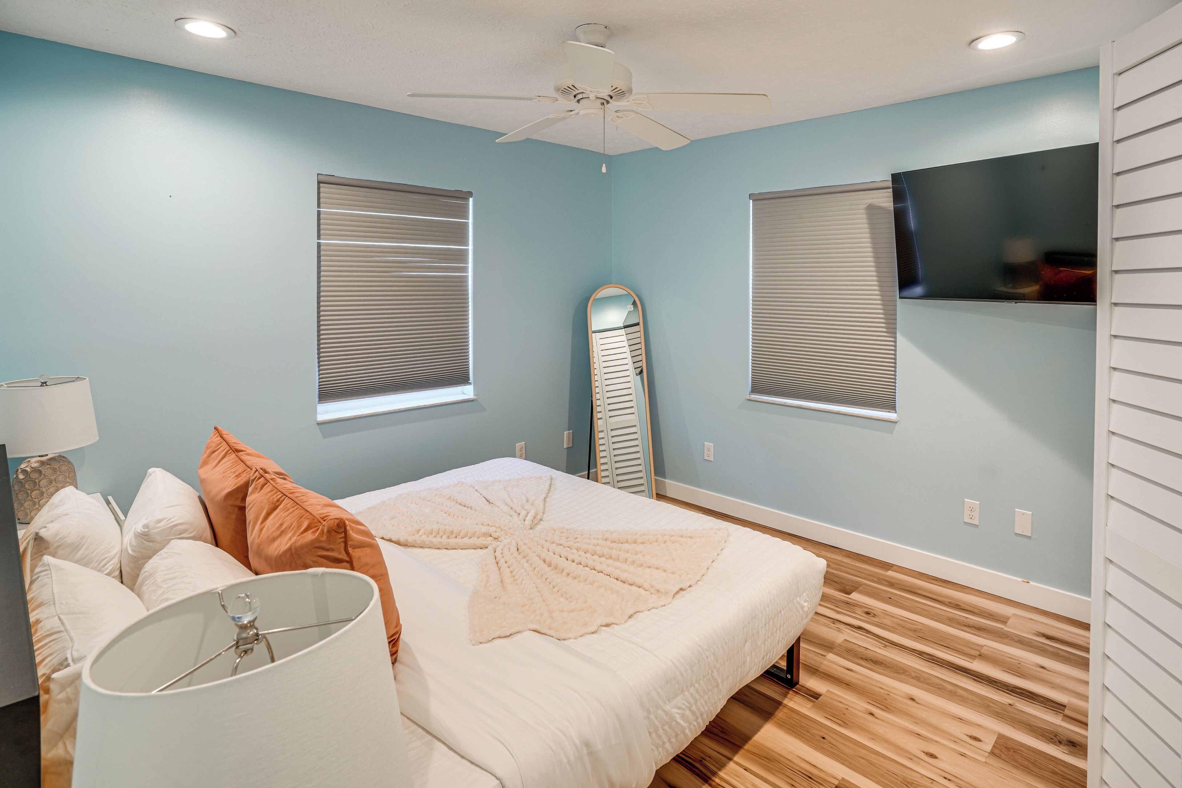 A bedroom with a bed, pillows, a mirror, a ceiling fan, and two windows with blinds.