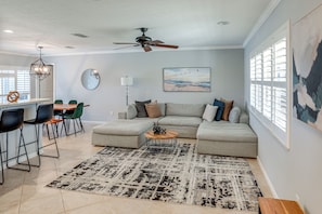 A modern living room with a sectional sofa, a dining area with chairs, a ceiling fan, and artwork on the walls.