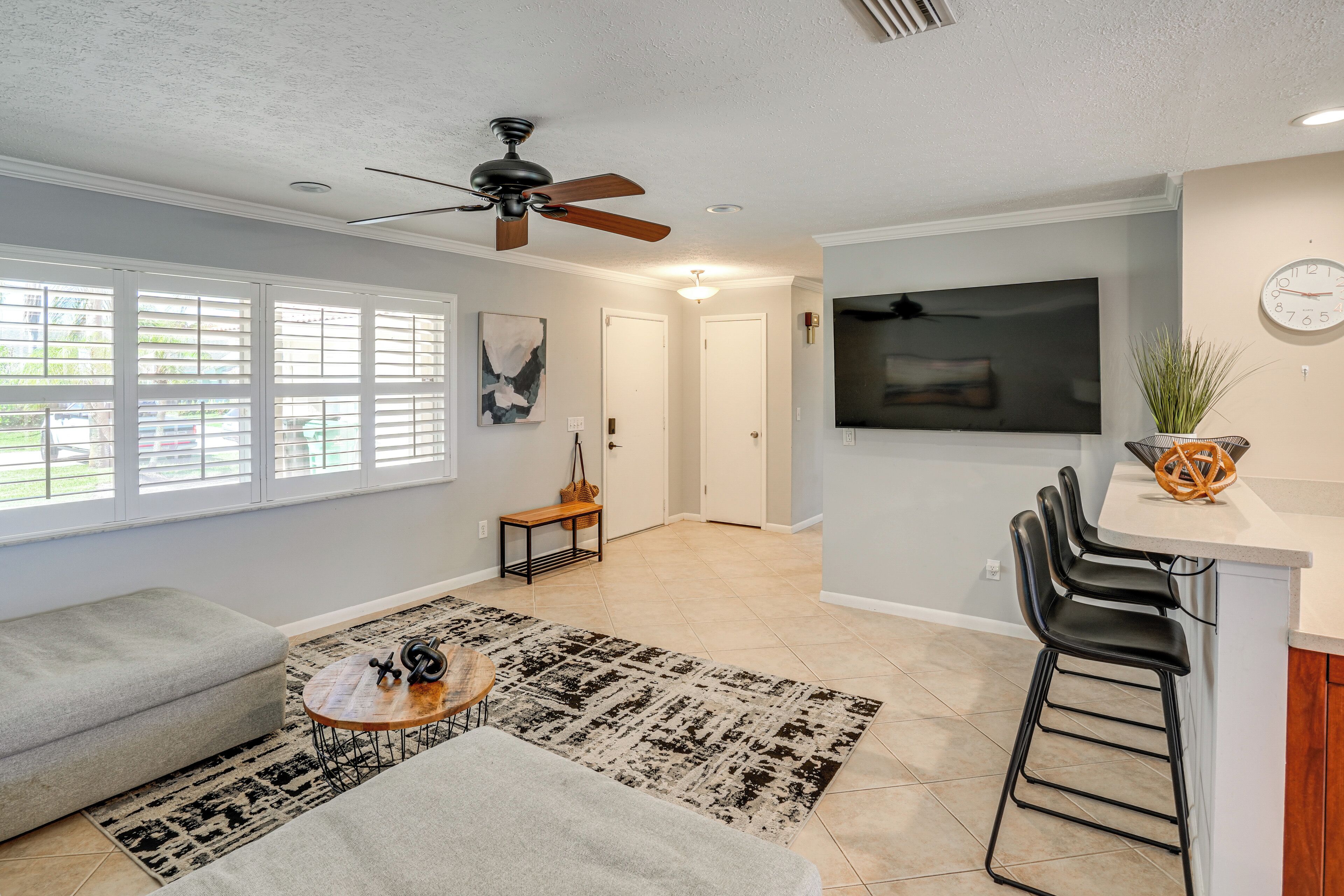 A modern living room with a sofa, coffee table, and a TV. There's a dining area with a bar and chairs.