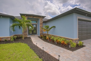 A modern two-story house with a stone facade, a garage, and a landscaped front yard with palm trees and a paved walkway. - Poolside Paradise in Northwest Cape Coral! (Cape Coral)