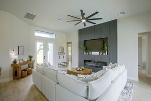 A modern living room with a sectional sofa, a ceiling fan, a flat-screen TV mounted on a wall, and a wooden coffee table. - Poolside Paradise in Northwest Cape Coral! (Cape Coral)