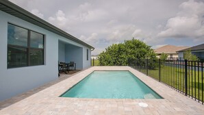 Ein Schwimmbecken mit Backsteinrand, eine Terrassenfläche mit Tisch und Stühlen sowie ein Sicherheitshinweisschild an einem Fenster.