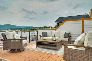 A deck with wicker furniture, a table, and a view of the water and mountains. - Brand-new Luxury Lakefront Condo - 2200 Sq Ft! (Lake George)